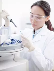 Scientist wearing lab coat, safety glasses, and gloves, holding pipette and vial next to centrifuge.