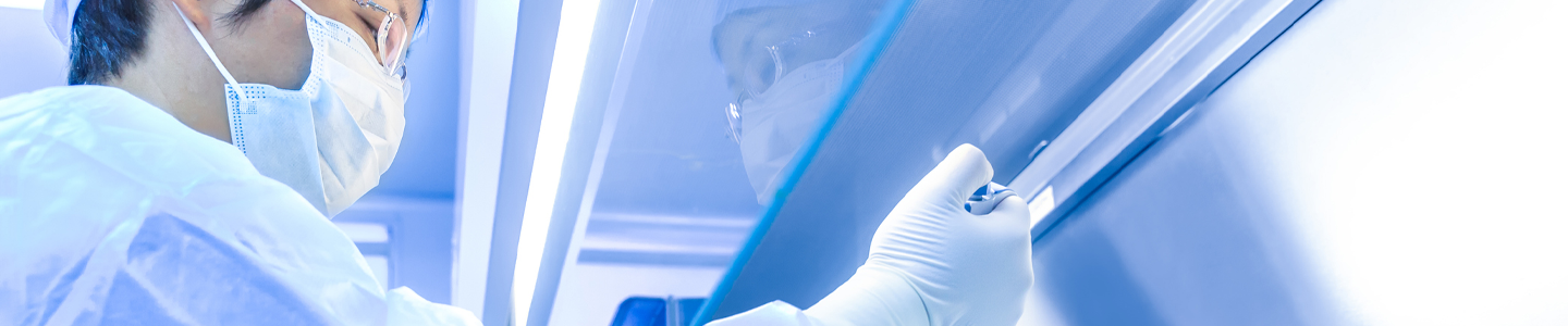 Scientist wearing cap, mask, gown, and gloves holding pipette and vial under a hood.