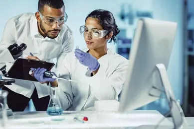 Two scientists wearing safety glasses looking at pipette and test tube and sitting in front of computer.