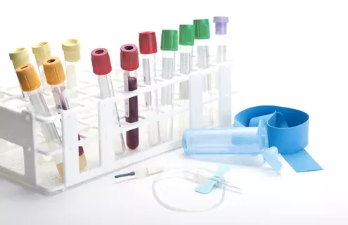 A collection of blood sample tubes in a white plastic rack, each with different colored caps (yellow, red, green, and purple). In front of the rack are medical tools including a blue tourniquet, a needle with tubing, and a clear plastic holder.