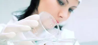 Female scientist wearing lab coat and gloves pouring clear liquid from beaker into funnel.
