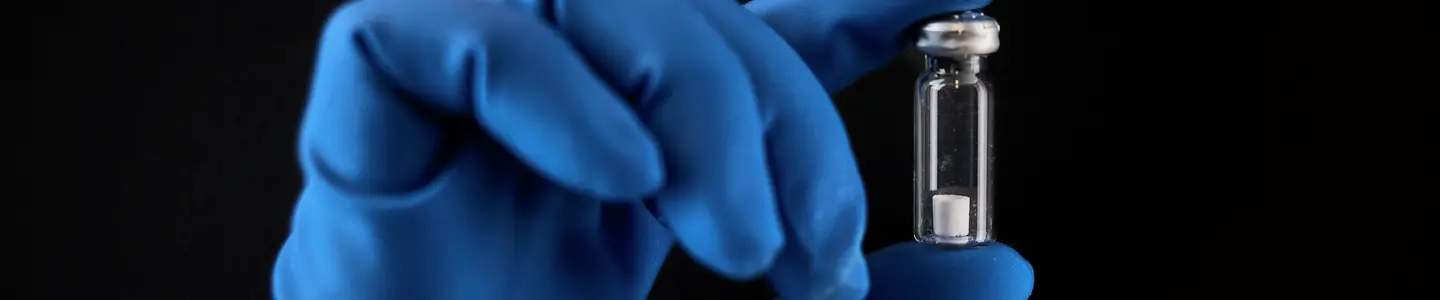 Blue gloved hand holding a Microquant vial between forefinger and thumb on a black background