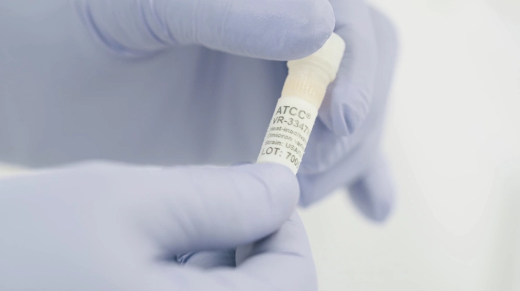 A gloved hand holds a labeled vial containing a viral strain sample, with "ATCC VR-3347" and "Omicron variant" visible on the label.