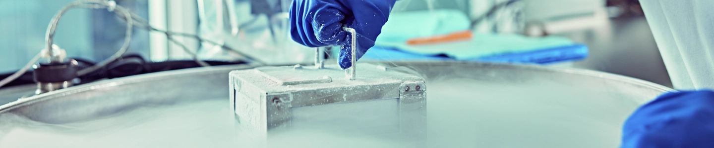 A scientist wearing blue gloves uses handle on a metallic container surrounded by vapor from liquid nitrogen, in a laboratory setting.