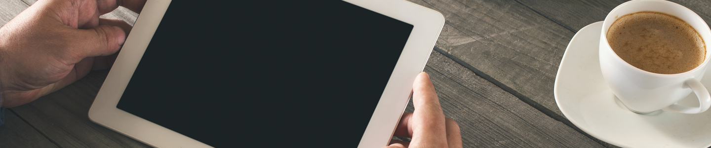 Hands holding a tablet on a wooden table with a cup of coffee.