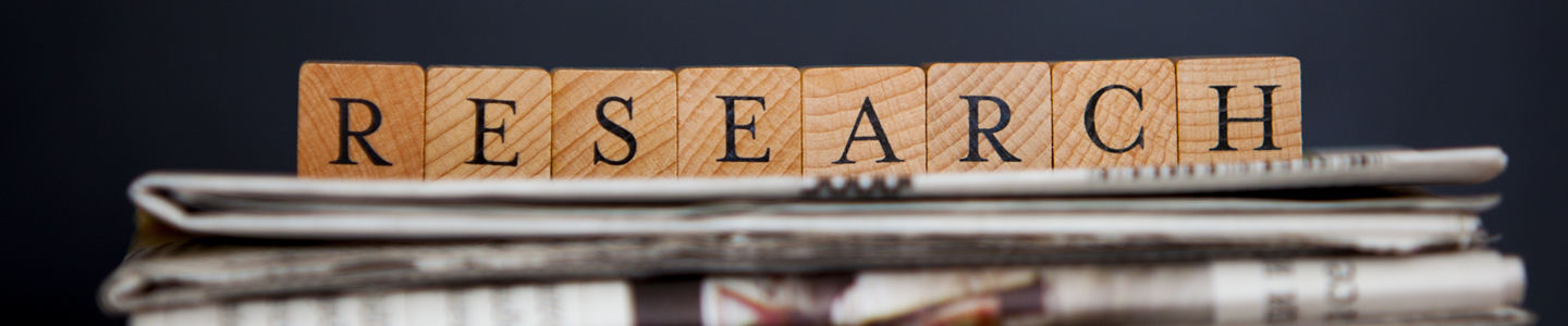 A row of small brown blocks, each with a letter, that spells "RESEARCH" on top of a stack of newspapers that appear blurry.