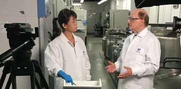 ATCC scientist with gloved hand in cooler on cart talking with Dr. Cypess, camera crew around them during filming in biorepository.