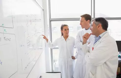 Three scientists in lab coats are standing in front of a whiteboard covered with complex diagrams and notes. One scientist is pointing at the whiteboard, explaining something to the others. The setting in a research facility with large windows in the background.