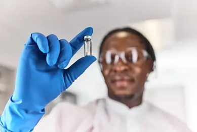Out of focus black, male, scientist wearing an in focus blue glove holding a Microquant vial to the left side of their face.