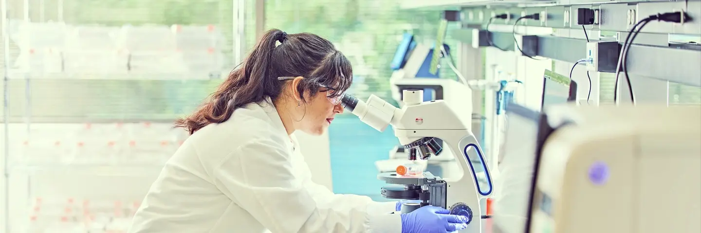 A scientist in a white lab coat and blue gloves looks through a microscope in a laboratory.
