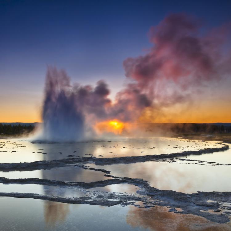 A plume of smoke rising from layers of slate-like liquid at Great Fountain Geyser in Yellowstone National Park at sunset.