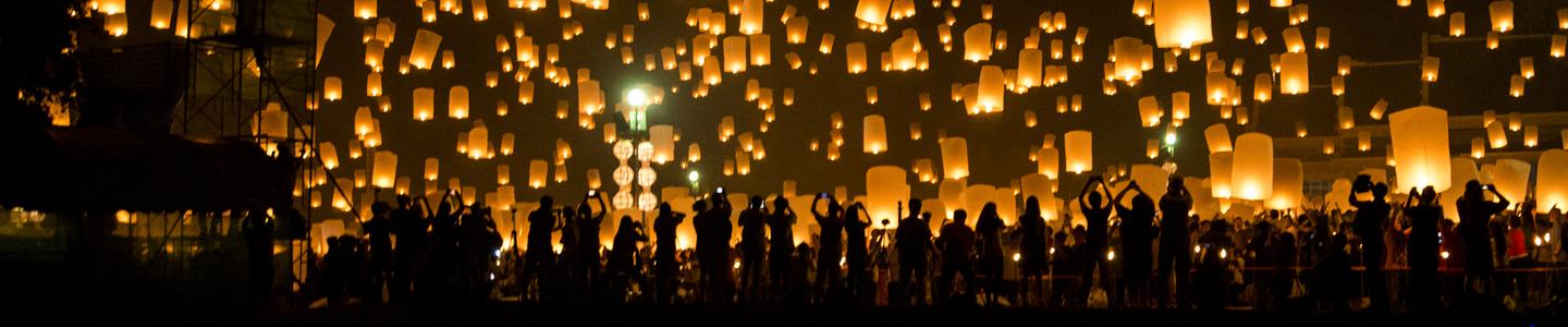 Silhouette of a crowd of people taking photos of amber-colored floating lanterns above their heads with the reflection in water behind them.
