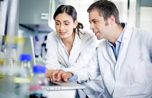 Female and male scientist in lab coats looking at laptop in a lab.