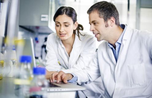 Female and male scientist in lab coats looking at laptop in a lab.