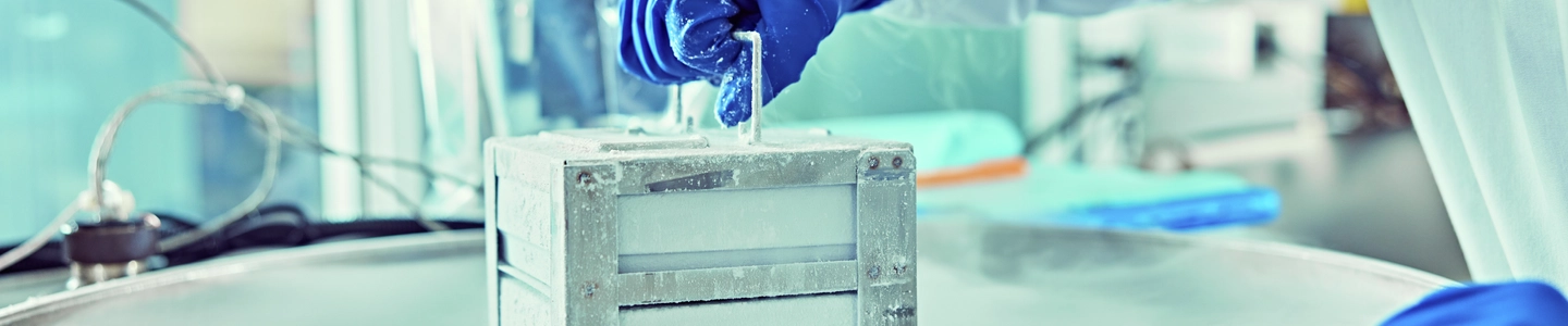 A scientist wearing blue gloves uses handle on a metallic container surrounded by vapor from liquid nitrogen, in a laboratory setting.