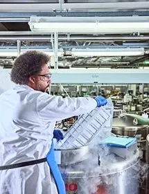 A laboratory technician in a white coat and blue gloves handles a tray of samples above a large stainless steel cryopreservation tank emitting vapor, likely from liquid nitrogen, with repository equipment visible in the background.
