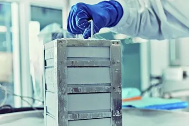 A scientist wearing blue gloves uses handle on a metallic container surrounded by vapor from liquid nitrogen, in a laboratory setting.