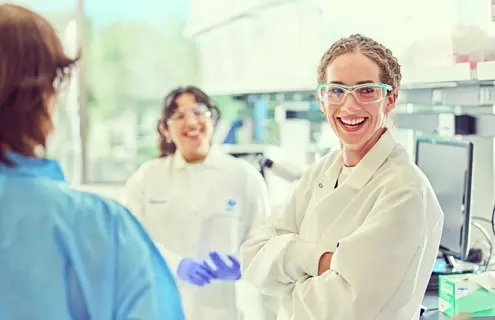 Three scientists in a laboratory setting, with one in the foreground wearing safety glasses and an ATCC lab coat, engaged in conversation near lab equipment including a microscope and computer.