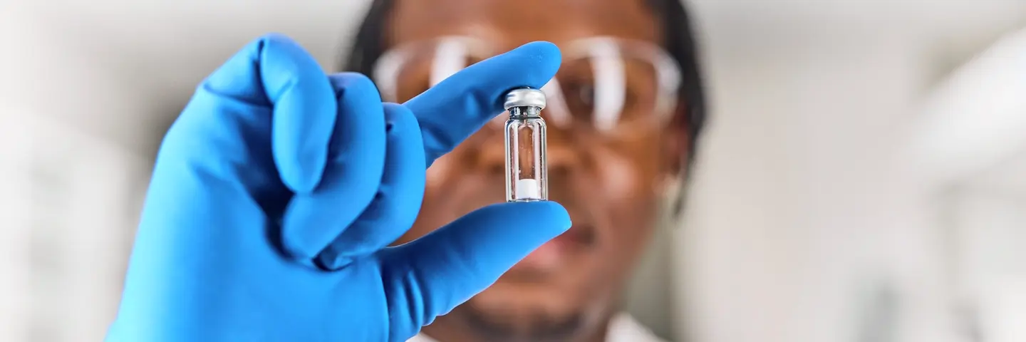 Out of focus black, male, scientist wearing a blue glove holding a Microquant vial infront of their face that is in focus. 