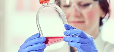 A scientist in blue gloves and a white lab coat holds up a t-flask with an orange cap, filled with red liquid. The background is blurred, showing shelves stocked with similar containers.