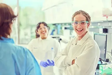 Three people in a lab, two in white coats and one in blue, with lab equipment in the background.