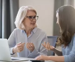 A gray-haired woman in glasses talking to a dark-haired woman at a desk in front of a laptop.