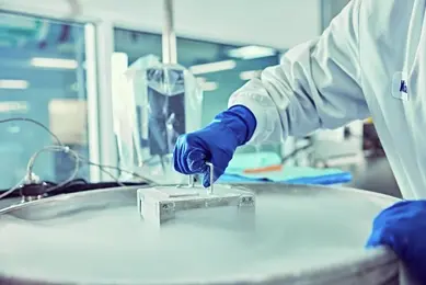 A scientist wearing blue gloves uses handle on a metallic container surrounded by vapor from liquid nitrogen, in a laboratory setting.