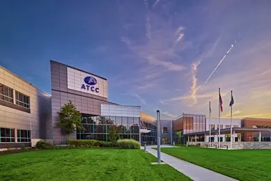 A wide exterior view of the ATCC campus at dusk shows a modern research facility with a glass‑fronted entrance, landscaped lawn, and a central walkway leading toward the main entrance. The ATCC logo is visible on the building facade beneath a softly lit evening sky.