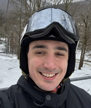 David Danko wearing a ski helmet and goggles on a snowy mountain chairlift, with leafless trees and ski trails in the background.
