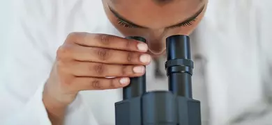 A person in a white lab coat adjusts the eyepiece of a black laboratory microscope, focusing on the instrument during use.