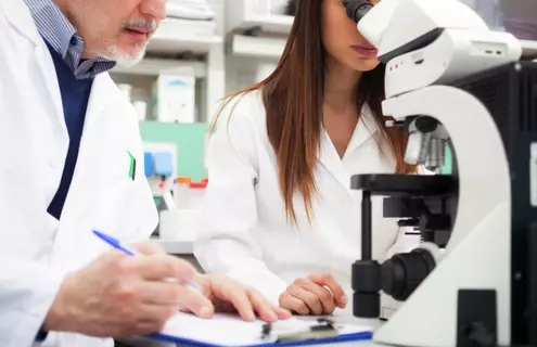 Scientist holding pen standing next to scientist looking into microscope.