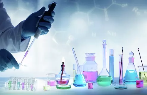 Table with flasks, beakers, and test tubes all holding different color liquids on top with a scientist on the left filling vials with a pipette.