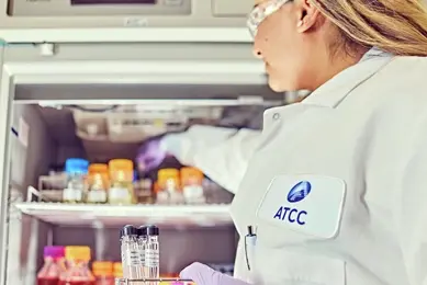 A scientist in an "ATCC"-labeled lab coat places a sample into a refrigerator stocked with vials and containers, holding a metal rack filled with test tubes.