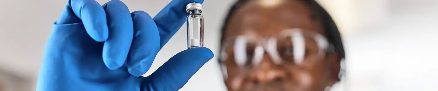 Out of focus black, male, scientist wearing an in focus blue glove holding a Microquant vial to the left side of their face.