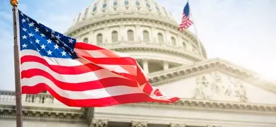 The image shows the United States Capitol building with a large American flag waving in the foreground and another American flag in the background.