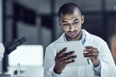 Male scientist in white lab coat, sitting next to microscope, looking at tablet.