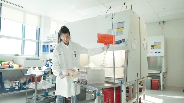 A laboratory technician in a white coat works under a biosafety cabinet, handling orange liquid-filled pipettes. The lab is well-lit with various equipment and supplies visible.