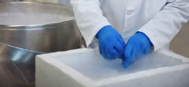 A person in a laboratory wearing a white lab coat labeled "Bobby" and "ATCC," handling an item inside a large container with blue gloves, surrounded by lab equipment.