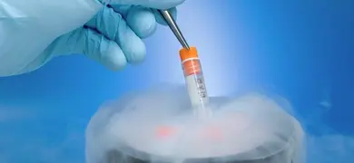 A gloved hand uses tweezers to lift a small vial with an orange cap from a container emitting dense white liquid nitrogen vapor.
