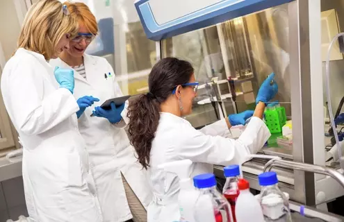 Two female scientists looking at tablet, standing behind seated female scientist who has gloved hands under a hood, holding a pipette.