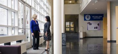 Two women in business attire standing inside office foyer near glass doors and windows.